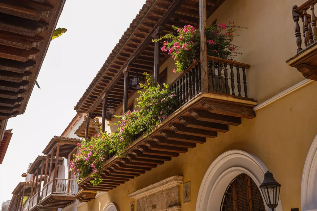 Balcón de madera con flores rosas y vegetación exuberante en un edificio colonial de color ocre.